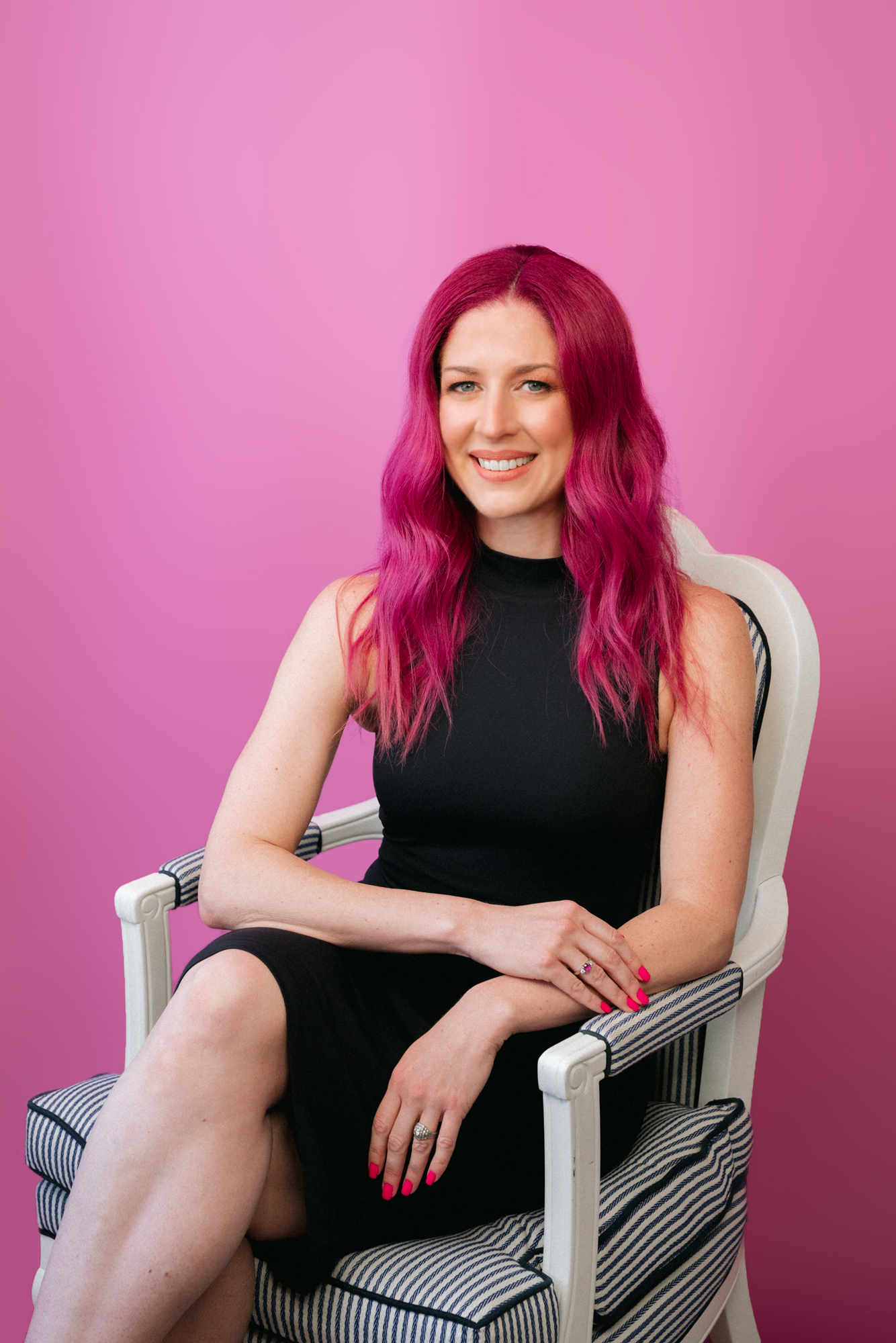 Woman with pink hair, wearing a black jacket and standing in front of a pink wall